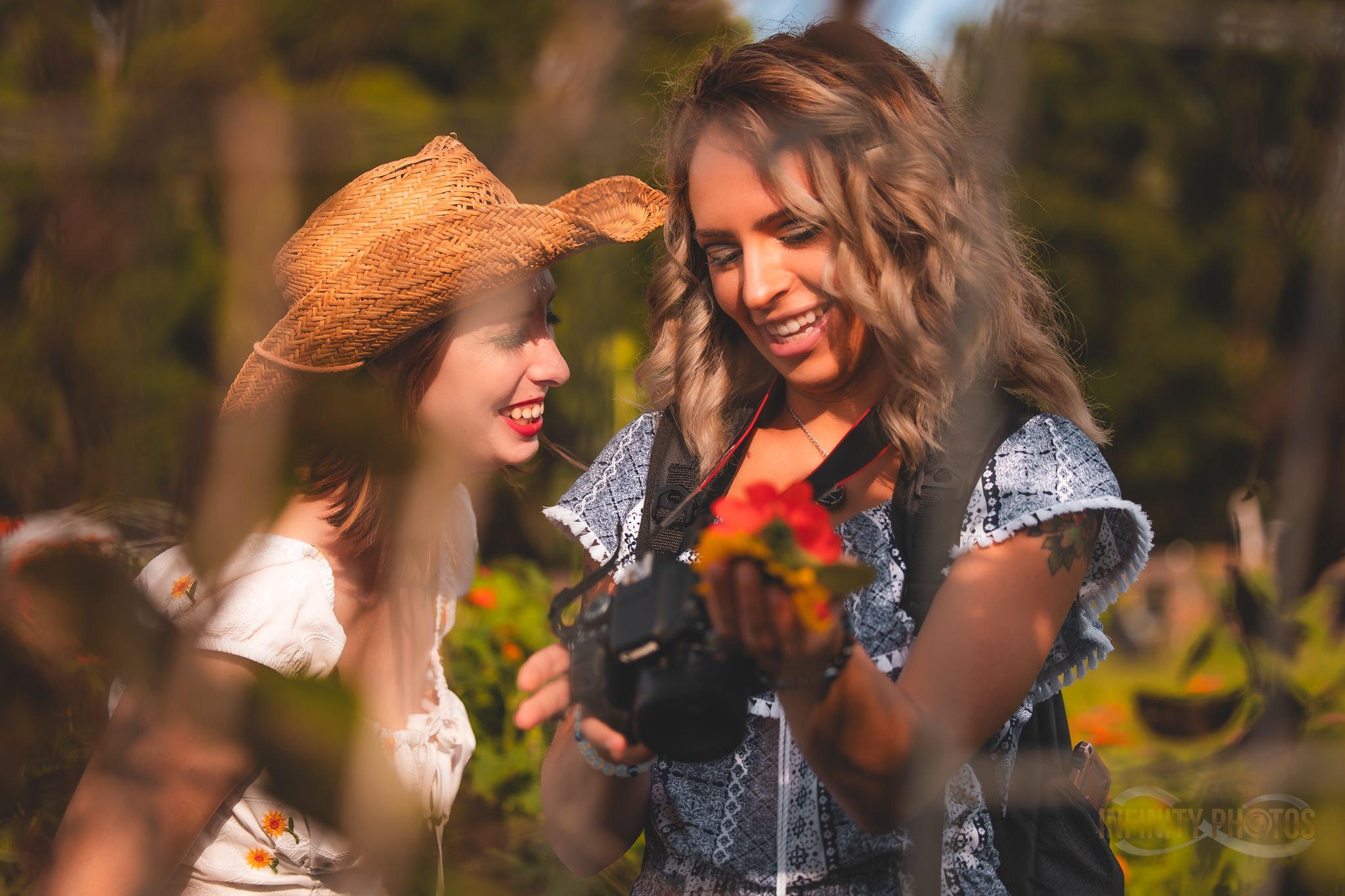 Two smiling women outdoors in natural light, one wearing a straw hat, admiring colorful flowers held in hand.