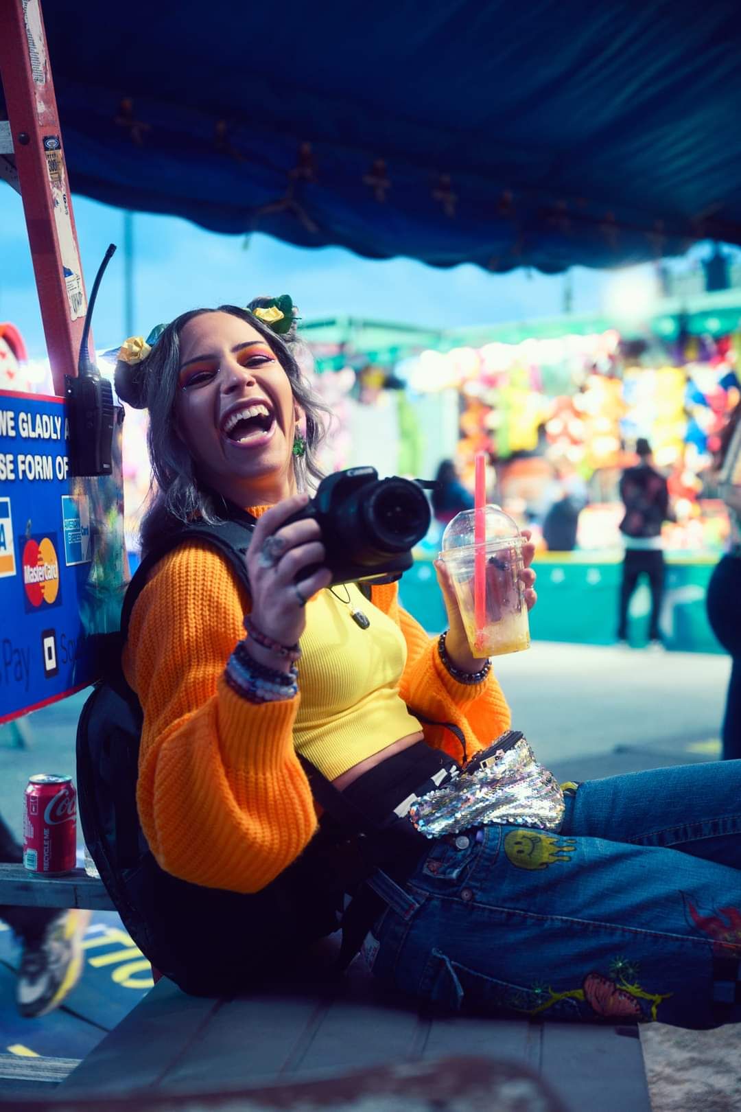 A laughing woman holds a DSLR camera and drink at a colorful outdoor carnival.