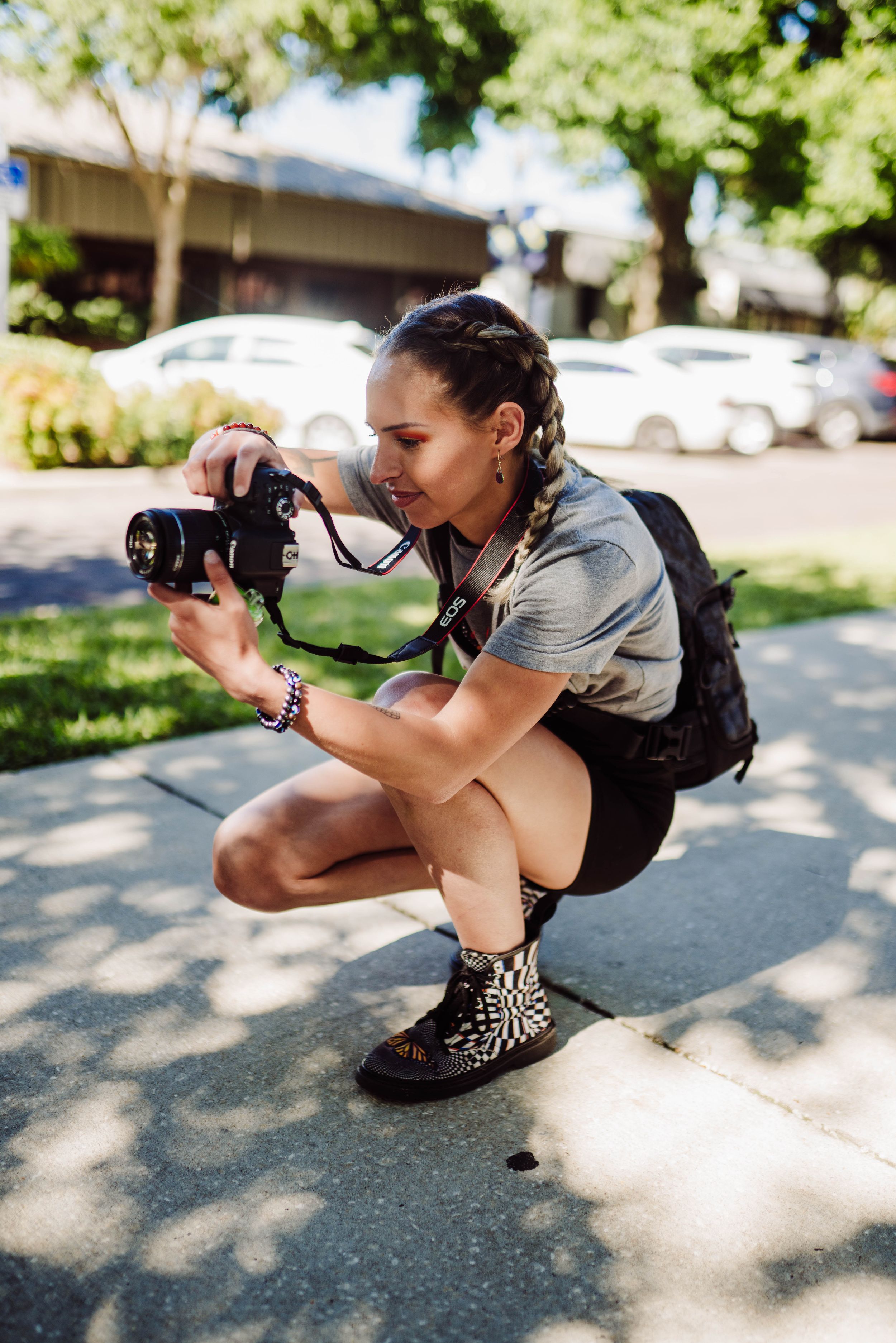 A young woman crouches on a sidewalk, carefully framing a shot with her DSLR camera.