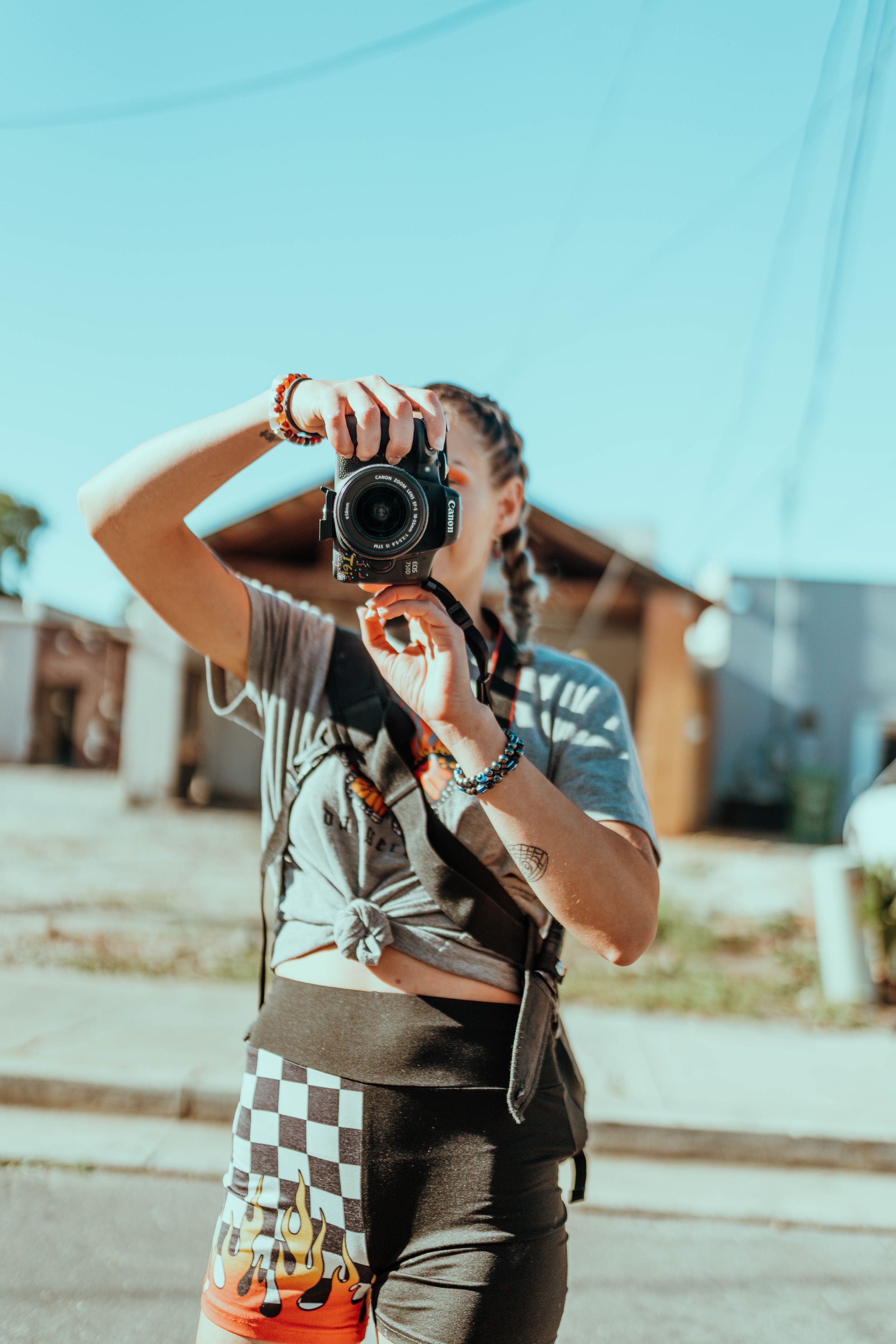 A woman with braids photographs outdoors on a sunny day using a DSLR camera.