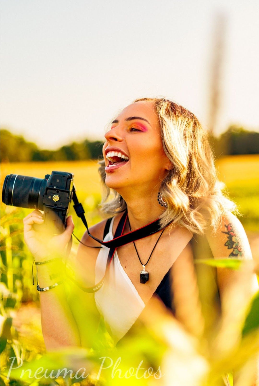 A joyful blonde woman laughs while holding a DSLR camera in a sunny field.