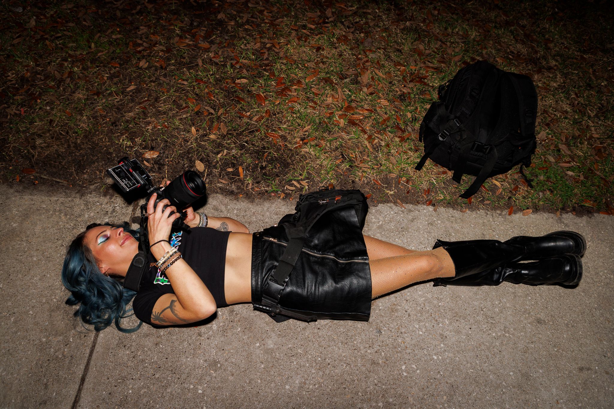 A woman in all-black outfit lies on pavement holding a camera at night.