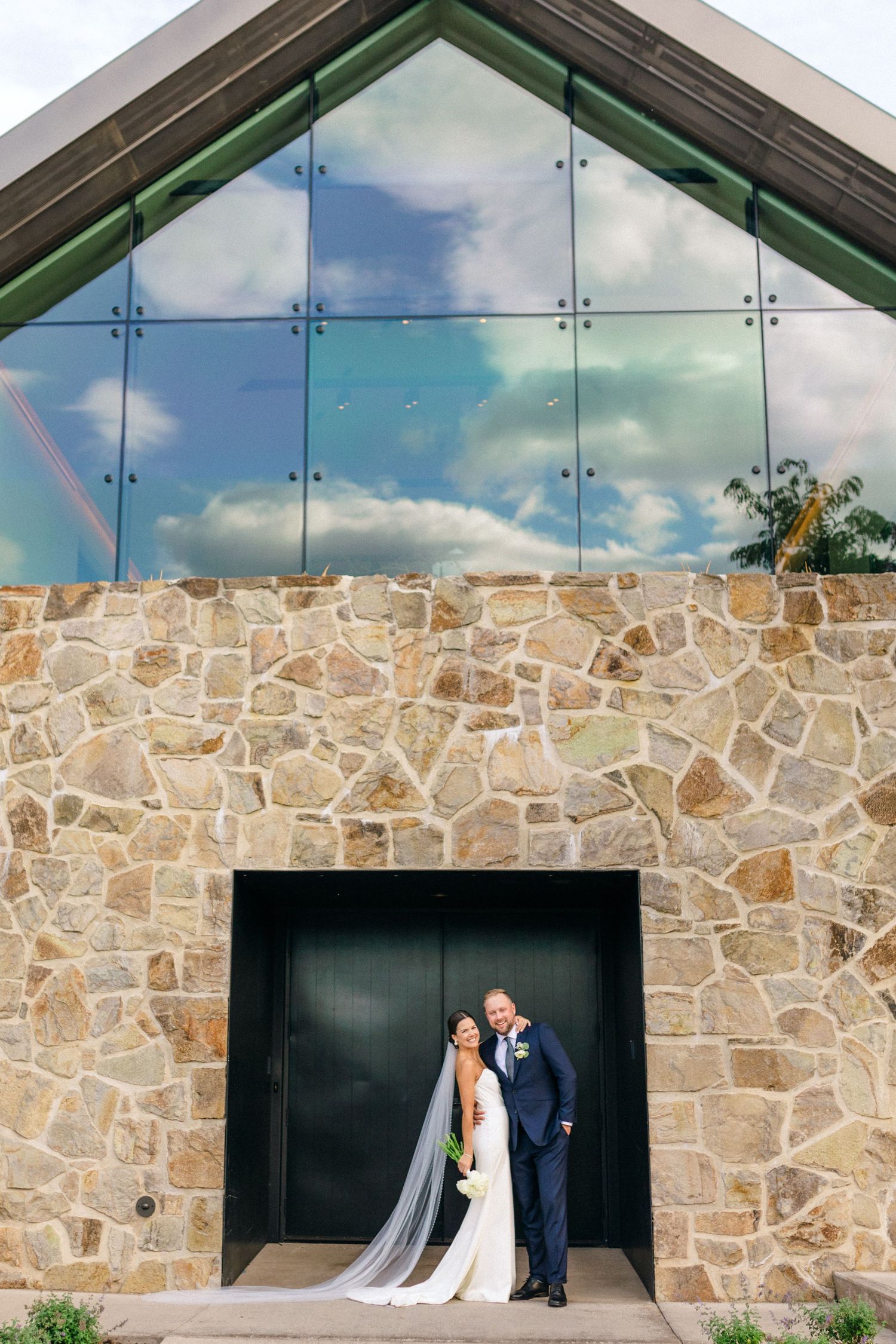 Couple in wedding attire stand in black doorway of stone church with glass windows reflecting clouds and blue sky above.