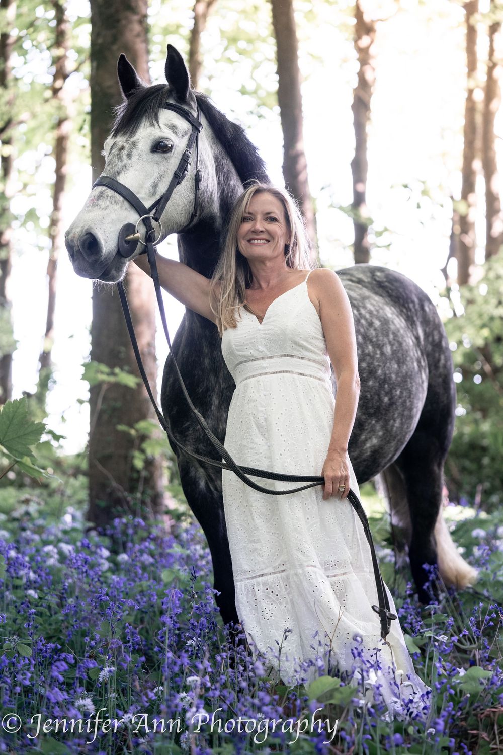 Grey dappled horse stands among blooming purple flowers in a forest setting.