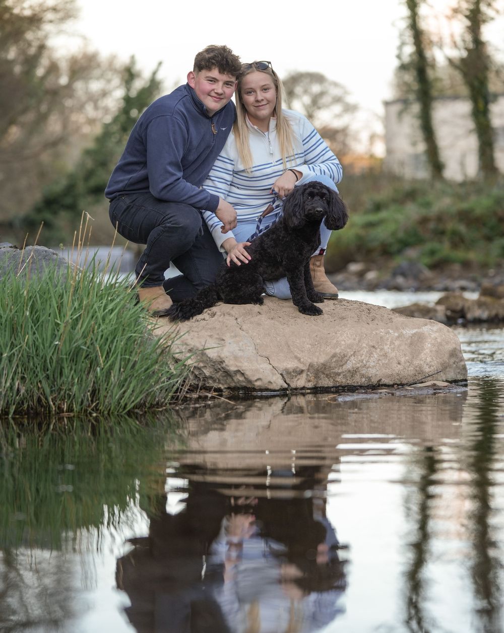 Couple and their black dog sitting by a reflective pond with nature backdrop.