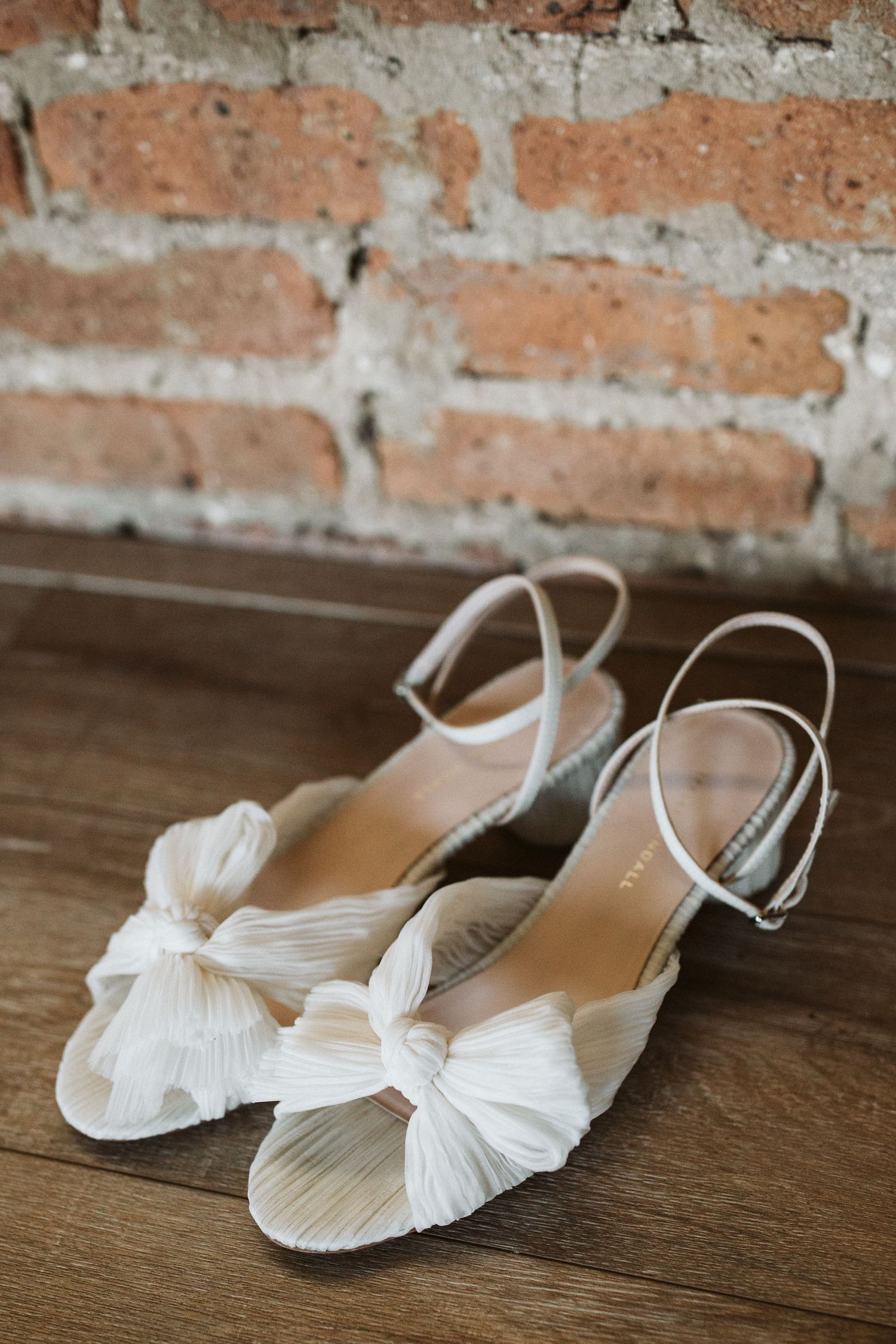 Elegant white bridal flats with bow details displayed on wooden flooring against brick wall.