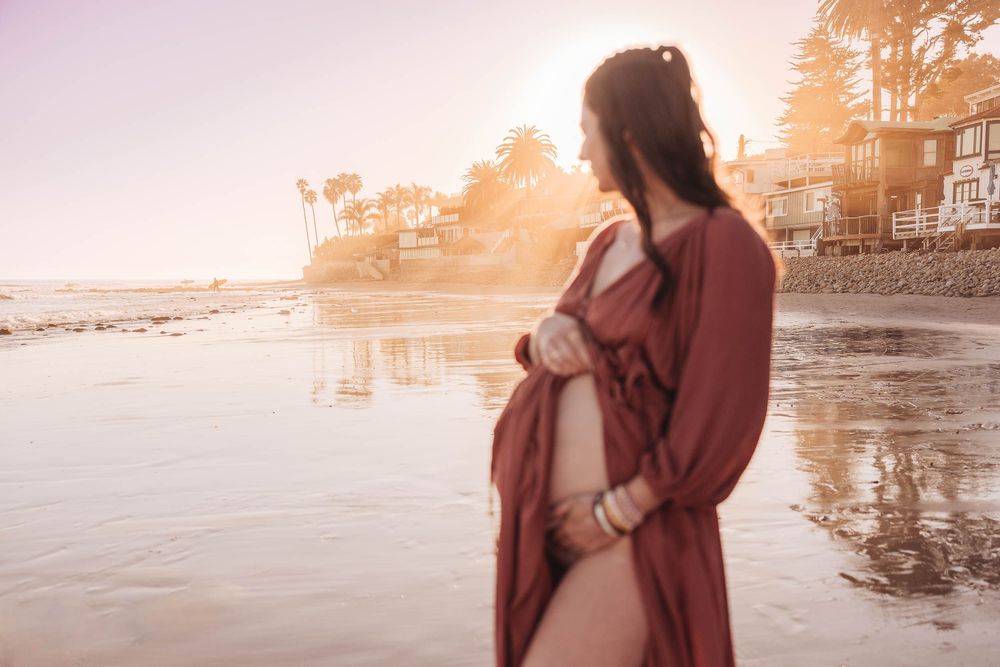 A person in a mauve dress stands on a beach at sunset with warm golden light and reflections on wet sand.