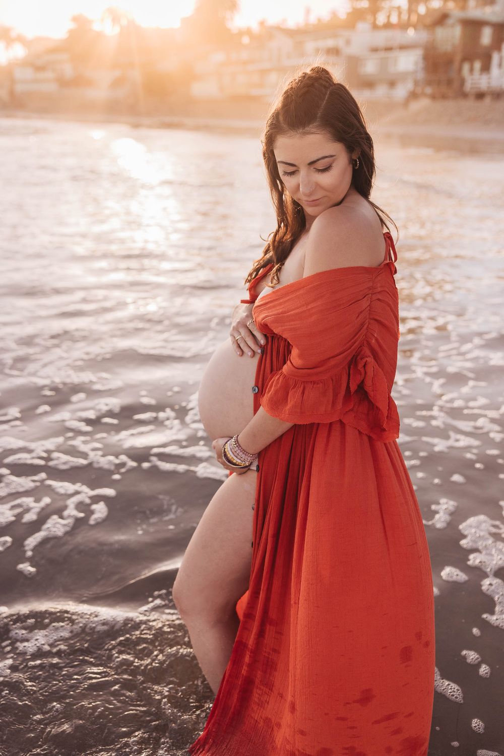 Woman in flowing red dress poses on beach during golden sunset with waves and coastline in background.