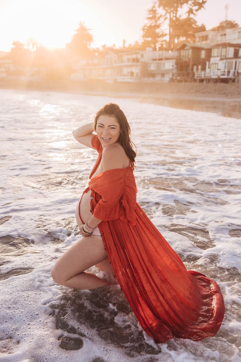 A person in a flowing red dress poses on the beach at sunset as ocean waves wash around them.