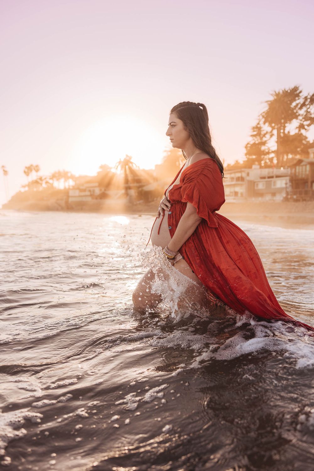 Woman in flowing red dress stands in ocean waves during dramatic sunset with beachfront homes visible.