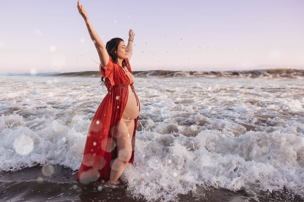 A person in a flowing red dress stands joyfully with arms raised as ocean waves crash around their feet at sunset.