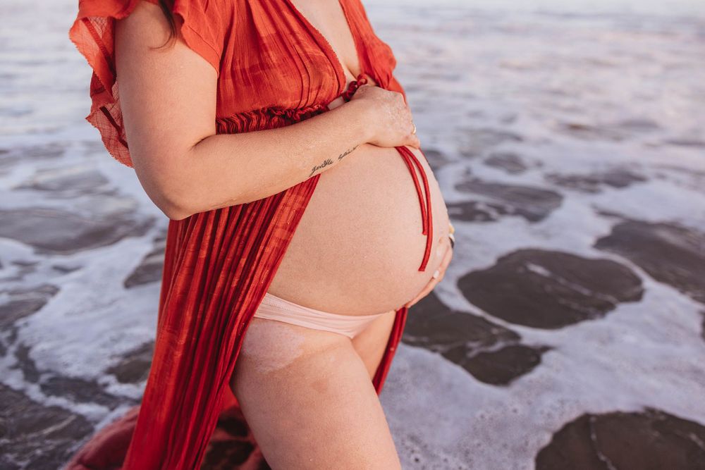 A pregnant figure in a flowing red dress poses at the beach with waves washing over dark sand at sunset.