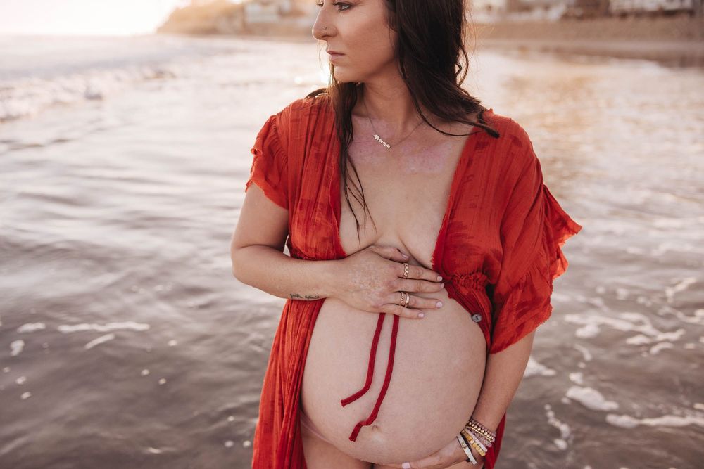Expecting woman wearing red shawl cradles her belly while standing on beach at sunset.