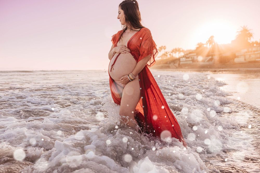 Expecting mother in flowing red dress stands in ocean waves during golden sunset photoshoot at the beach.