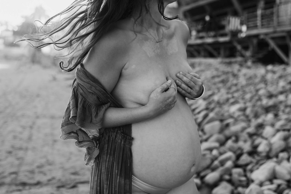 Black and white artistic photograph of a pregnant woman in a flowy dress standing on a rocky beach.