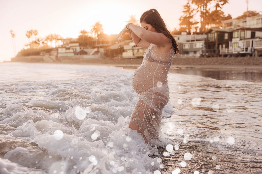 A person in a beige dress splashes through shallow ocean waves at sunset along a beach with houses in the background.