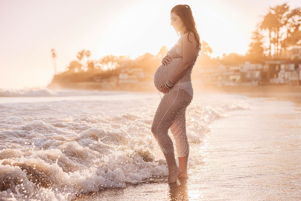 Silhouetted pregnant woman stands in ocean waves at sunset on tropical beach.