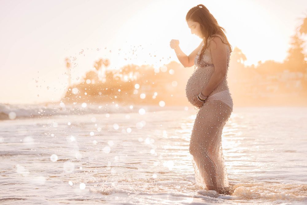 Silhouetted pregnant figure standing in ocean waves at sunset with water splashing around her.