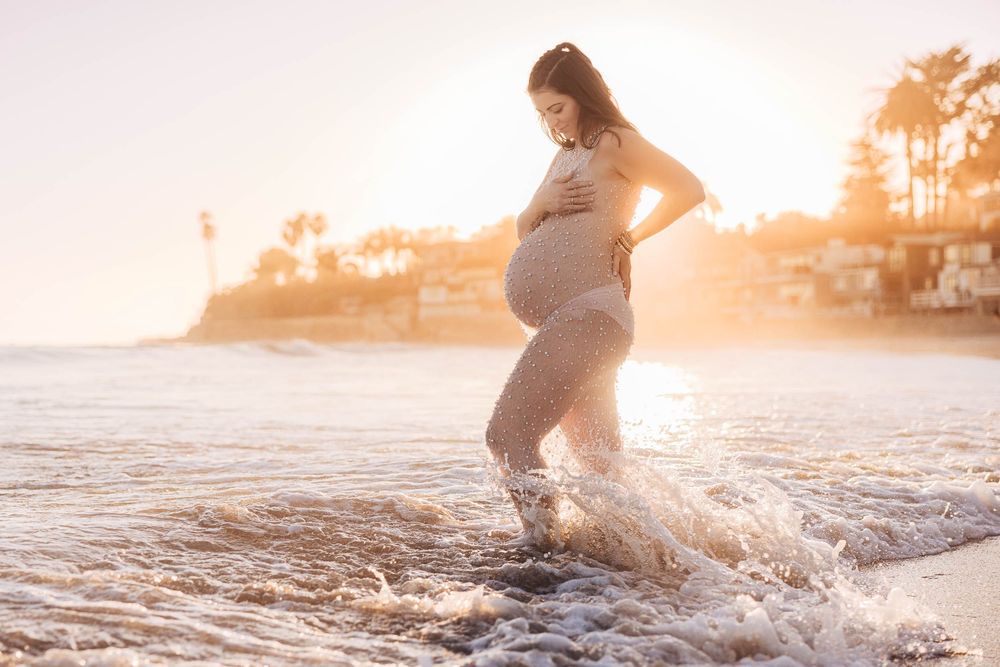 An expectant mother in a flowing dress stands in ocean waves at sunset, creating a dreamy silhouette against golden light.