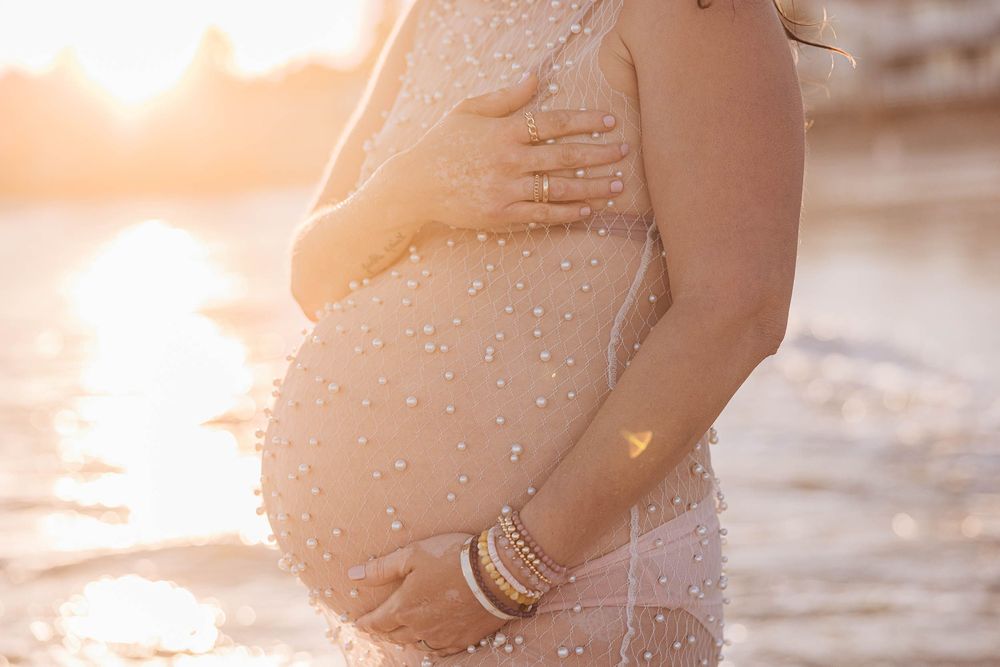 Silhouette of a pregnant woman's belly with sunlight streaming through water droplets at golden hour on the beach.