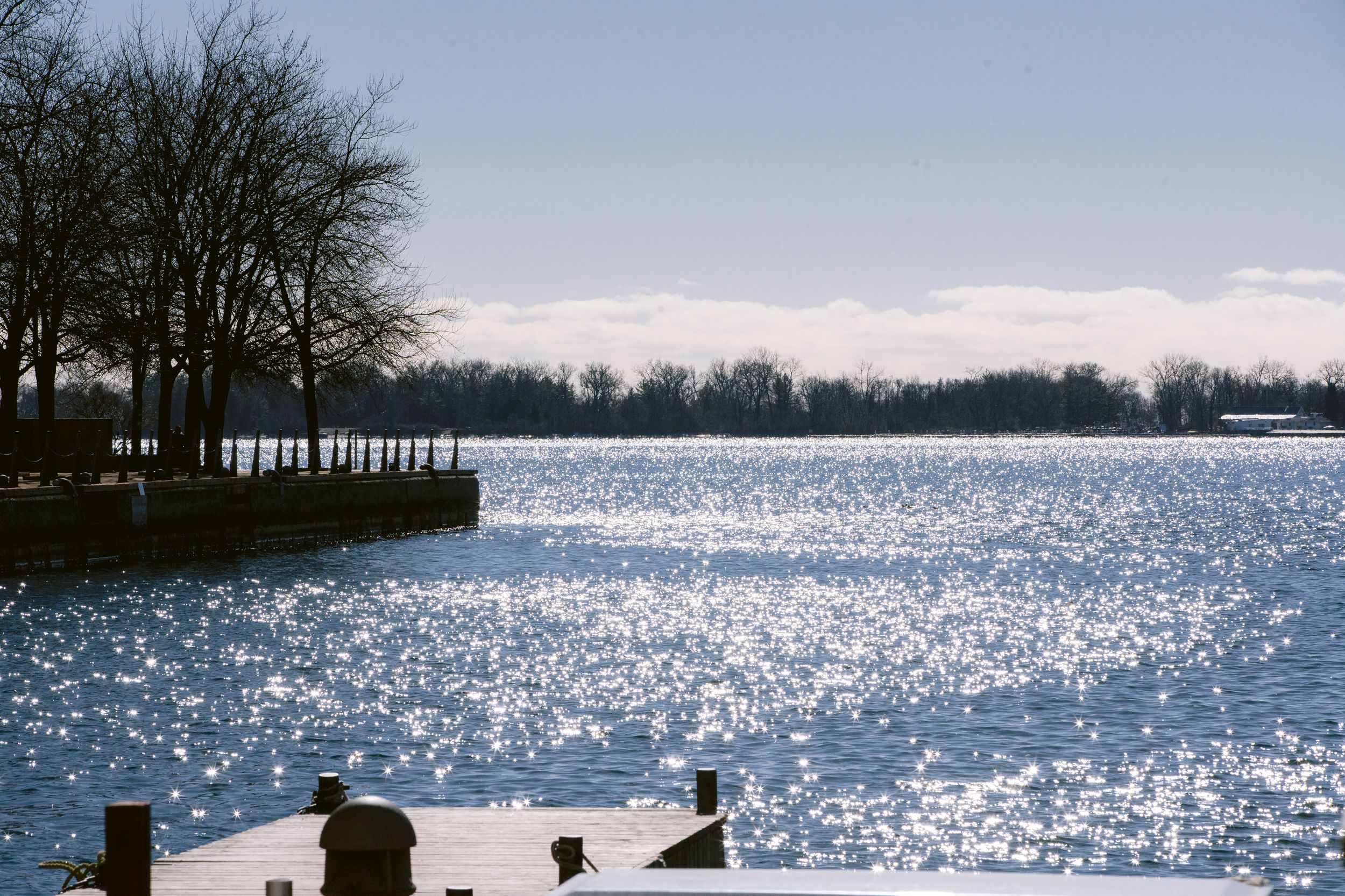 Sunlight sparkles across a winter lake with silhouetted trees along the shoreline and dark wooden posts in the foreground.