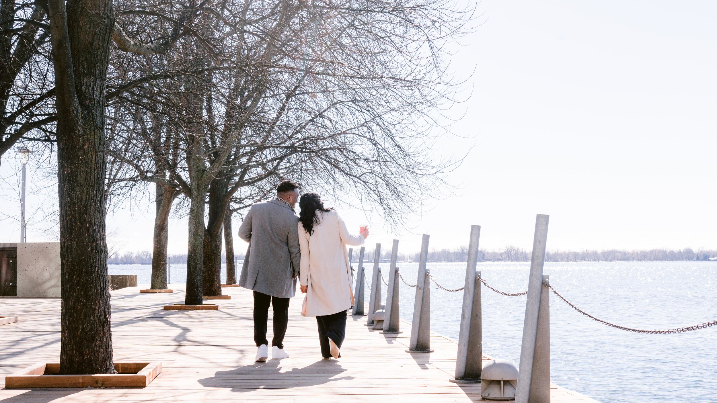 Two people walking along a snowy lakeside promenade with bare winter trees and white rope barriers.