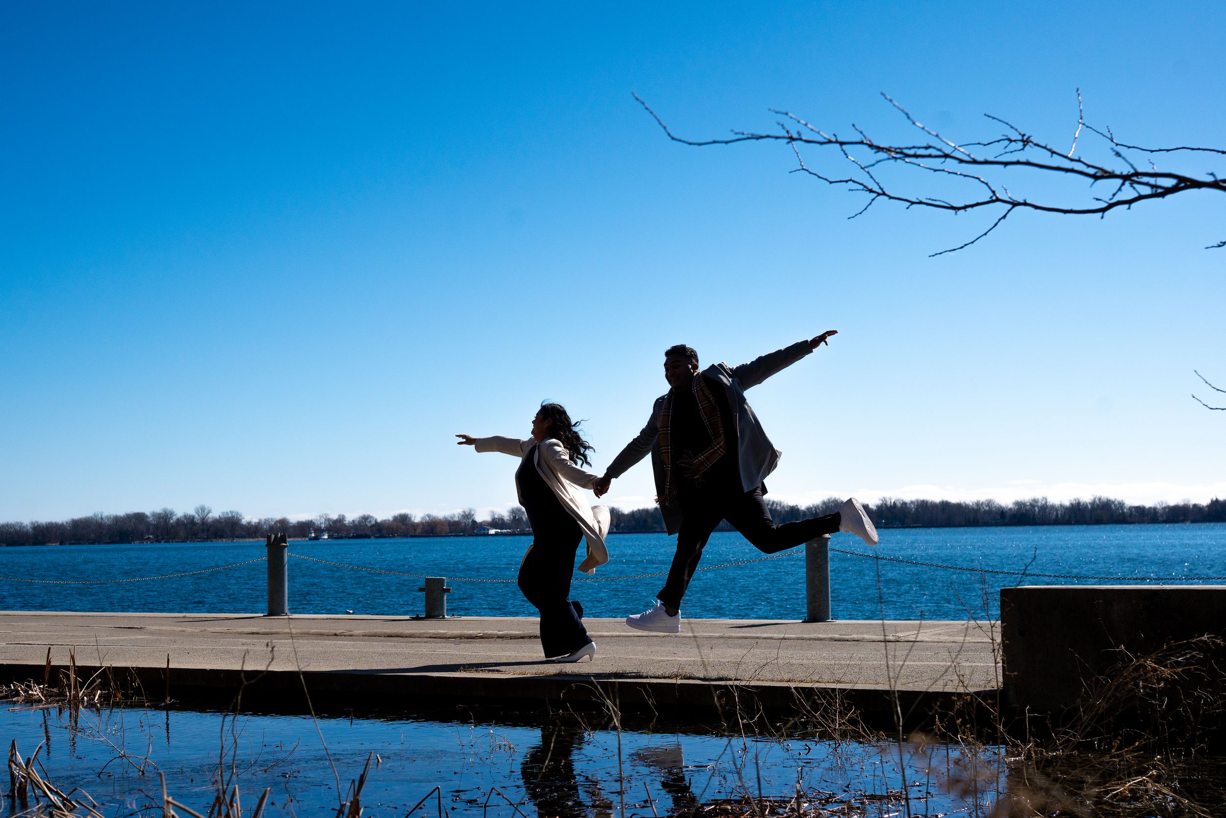 Silhouetted figures dancing playfully on a dock by a lake under bright blue sky.