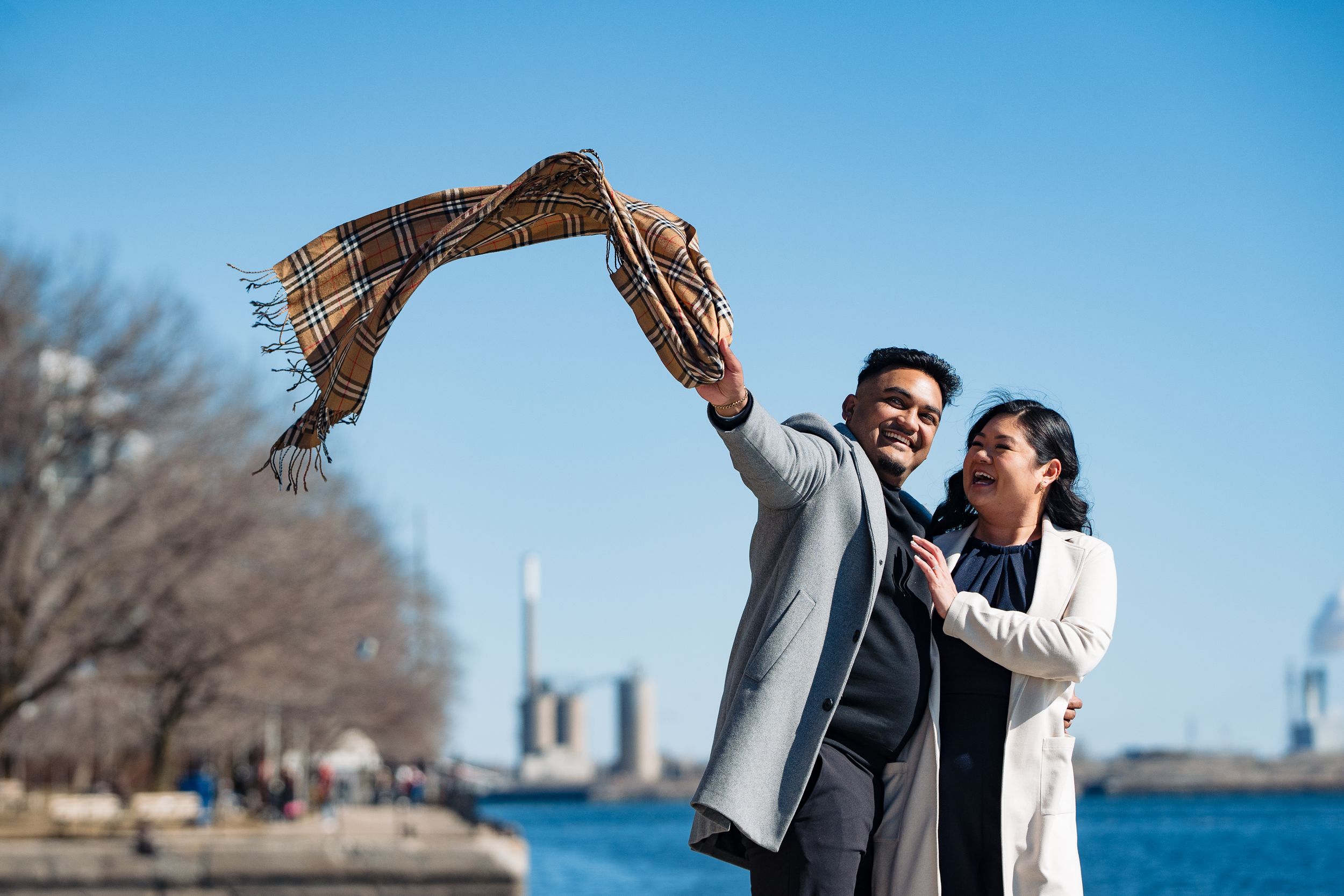 A couple enjoys a playful moment by the waterfront as they hold up a warm scarf against a bright blue sky and cityscape.