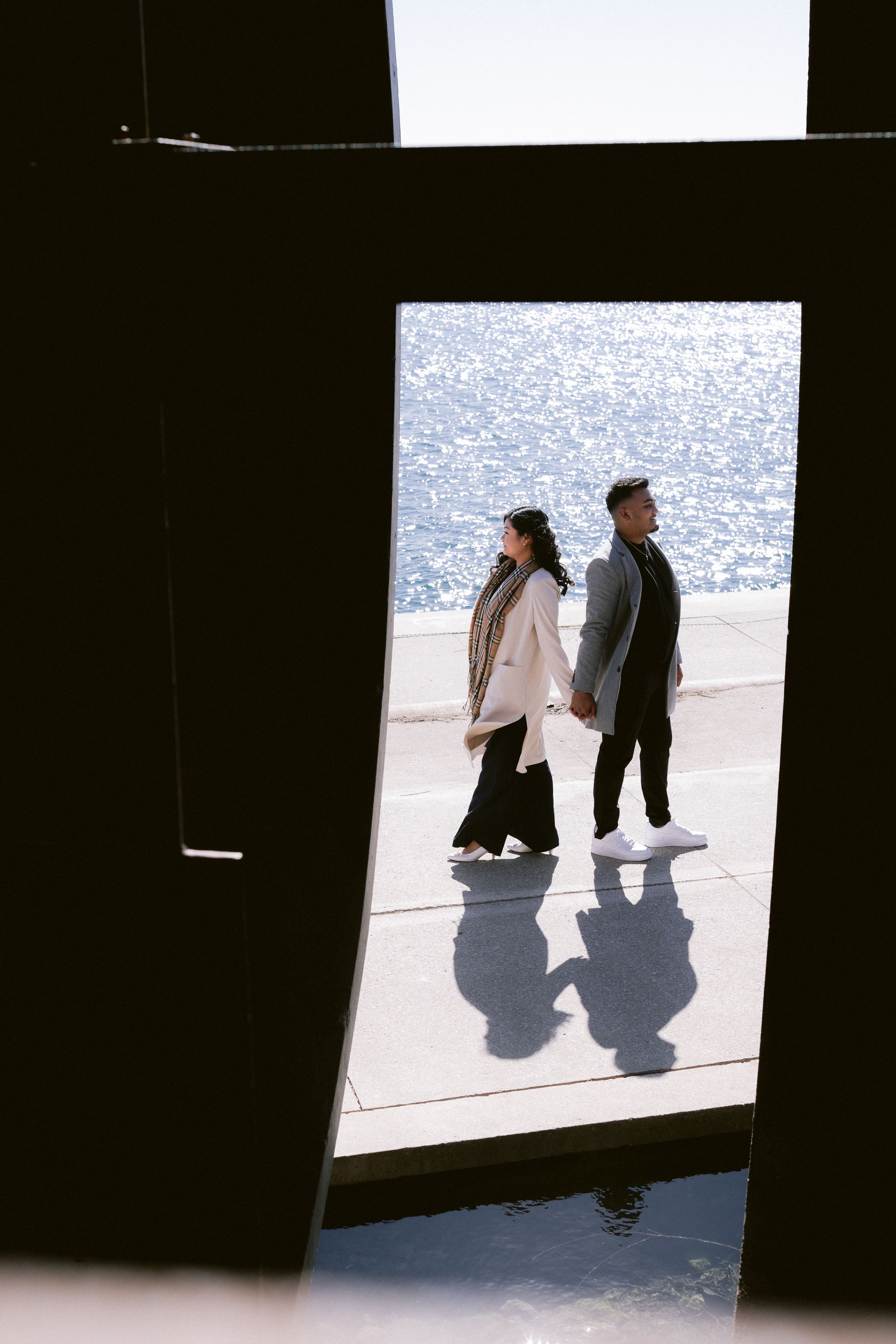 Artistic black and white photo of a couple walking along a waterfront, casting long shadows.