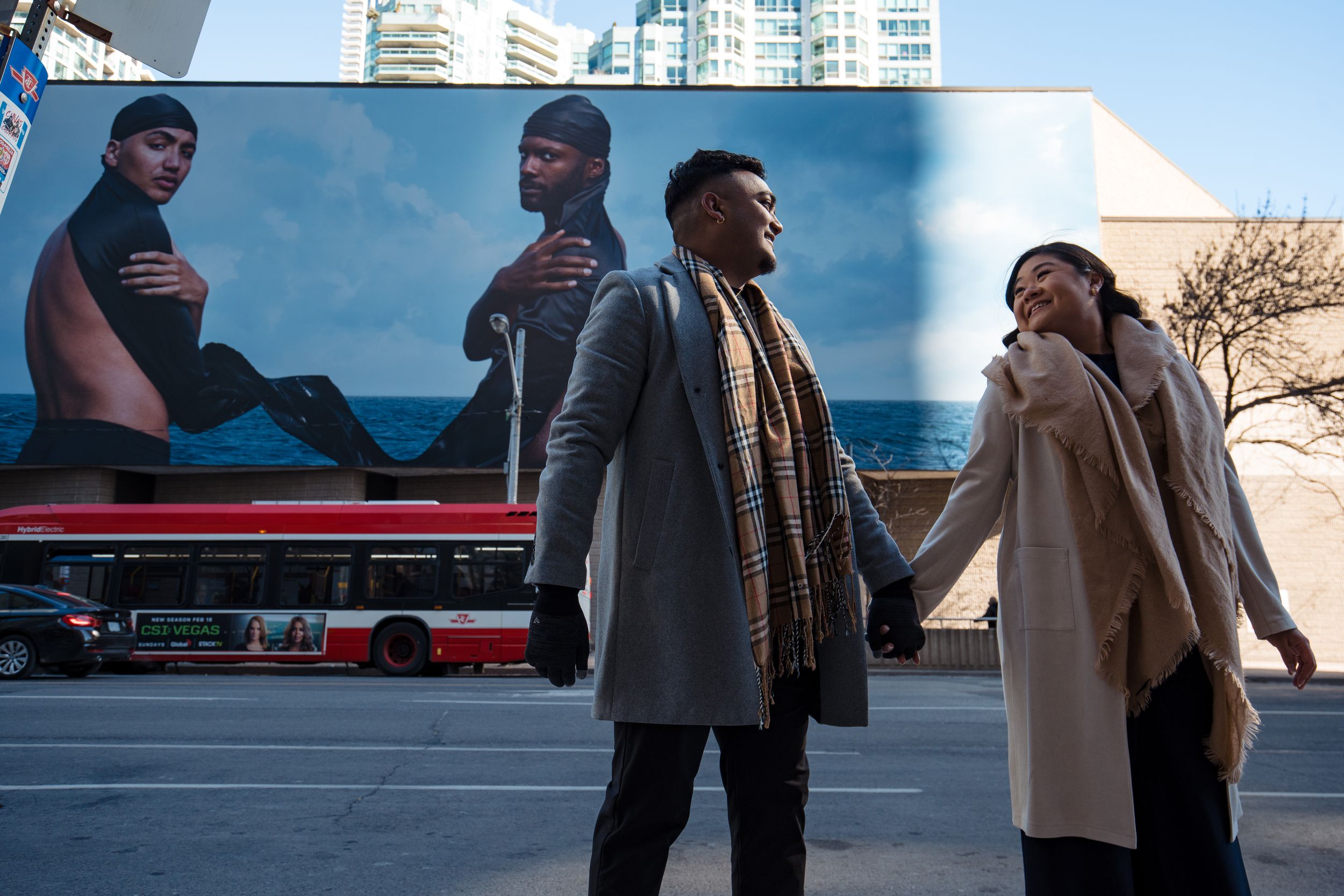 Two people in winter coats having a conversation on a city street with a large billboard and bus in the background.