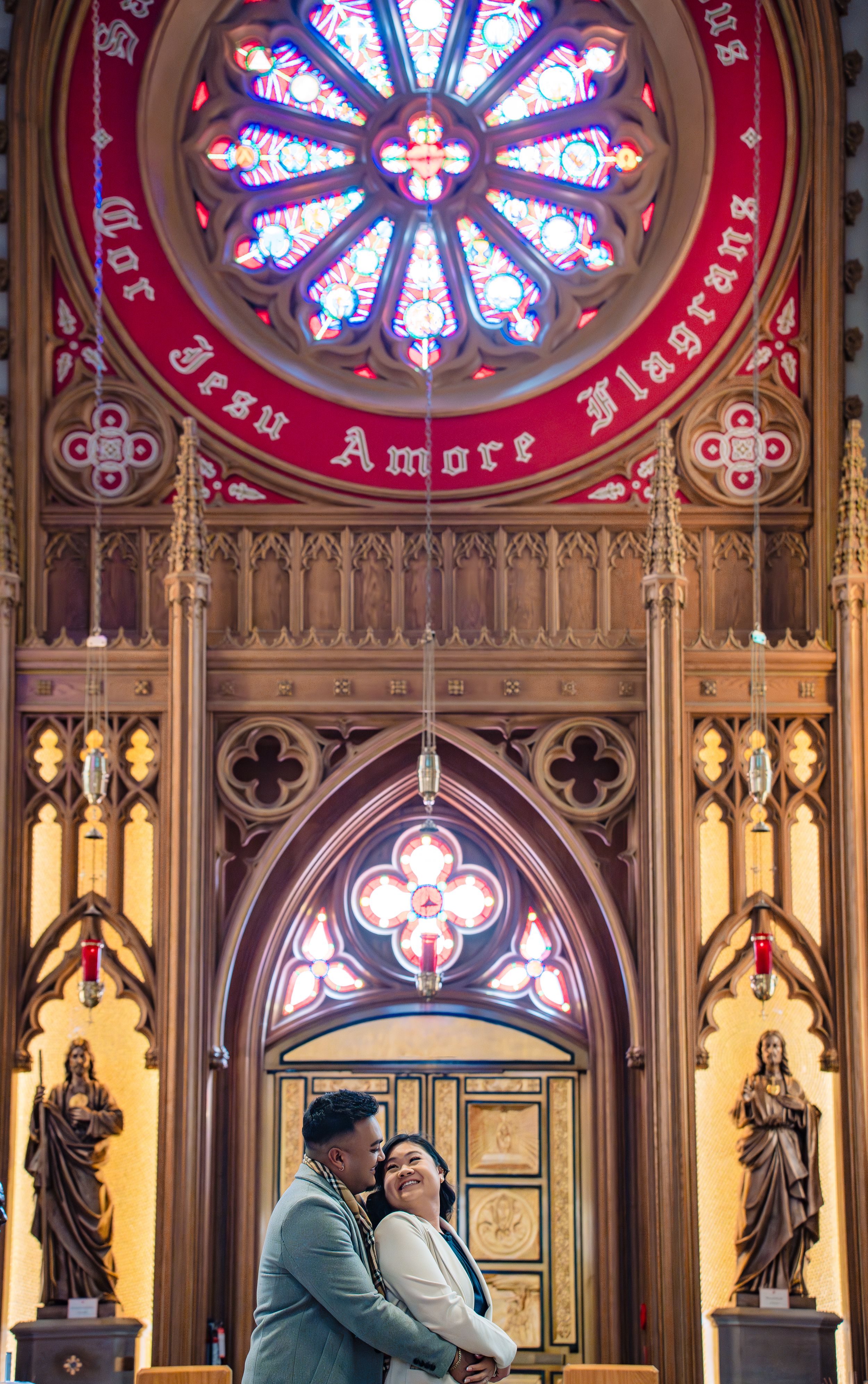 Ornate church interior featuring a rose stained glass window, gothic arches, and religious statues along stone walls.