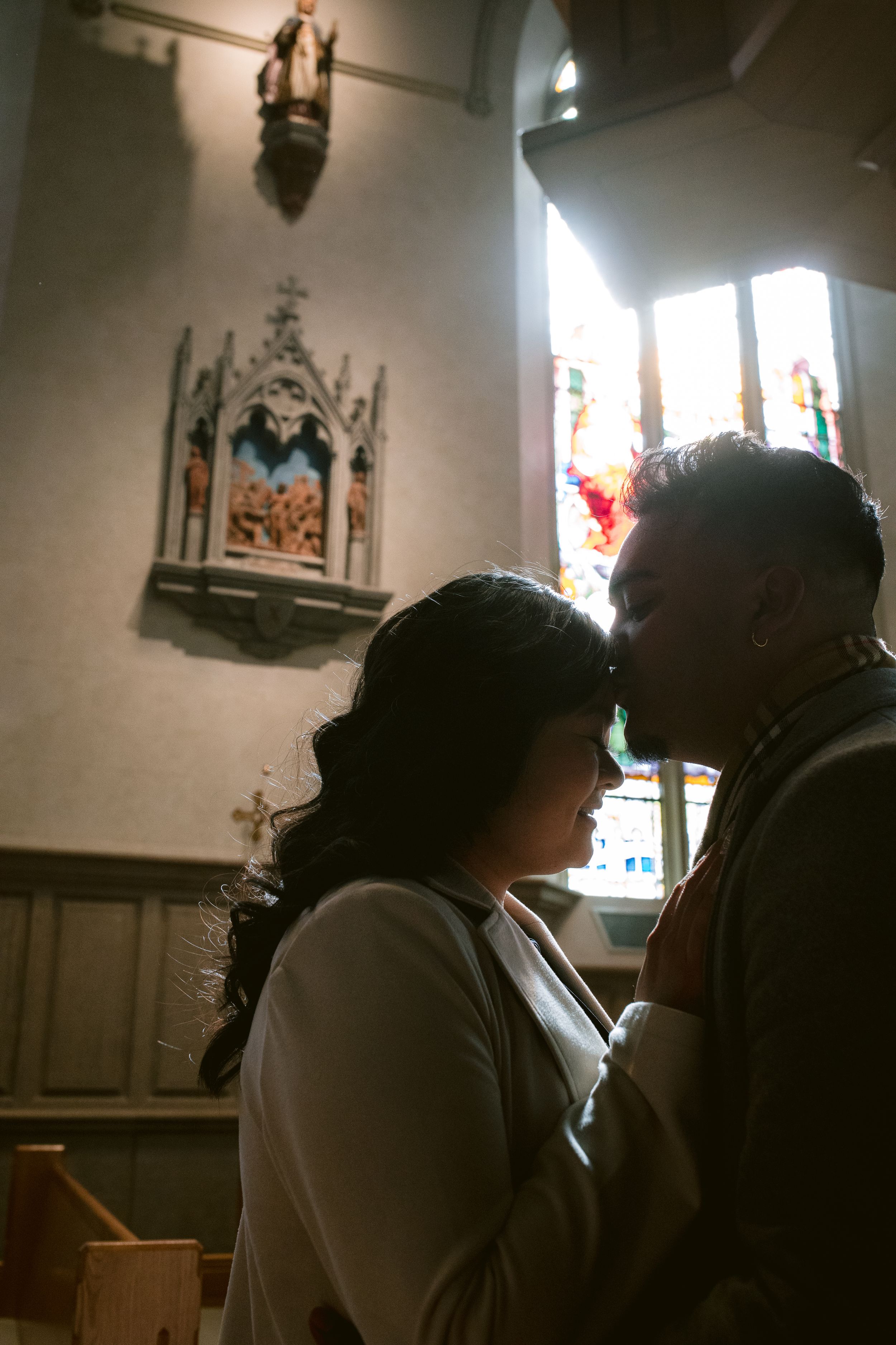 Silhouettes stand in intimate embrace inside a church with stained glass windows and religious artwork visible.