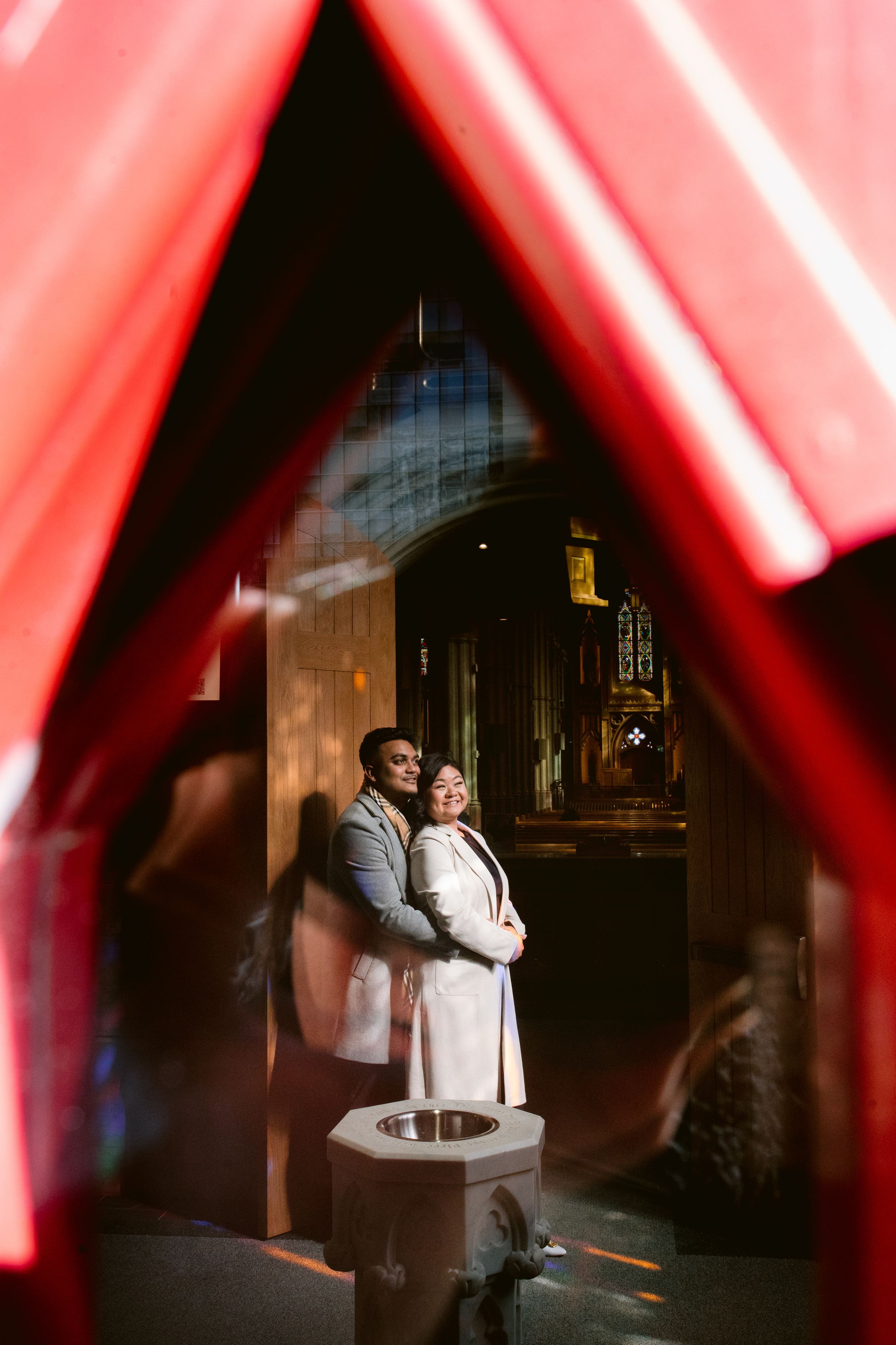 Couple posing under red architectural elements with dramatic lighting and gothic church background.