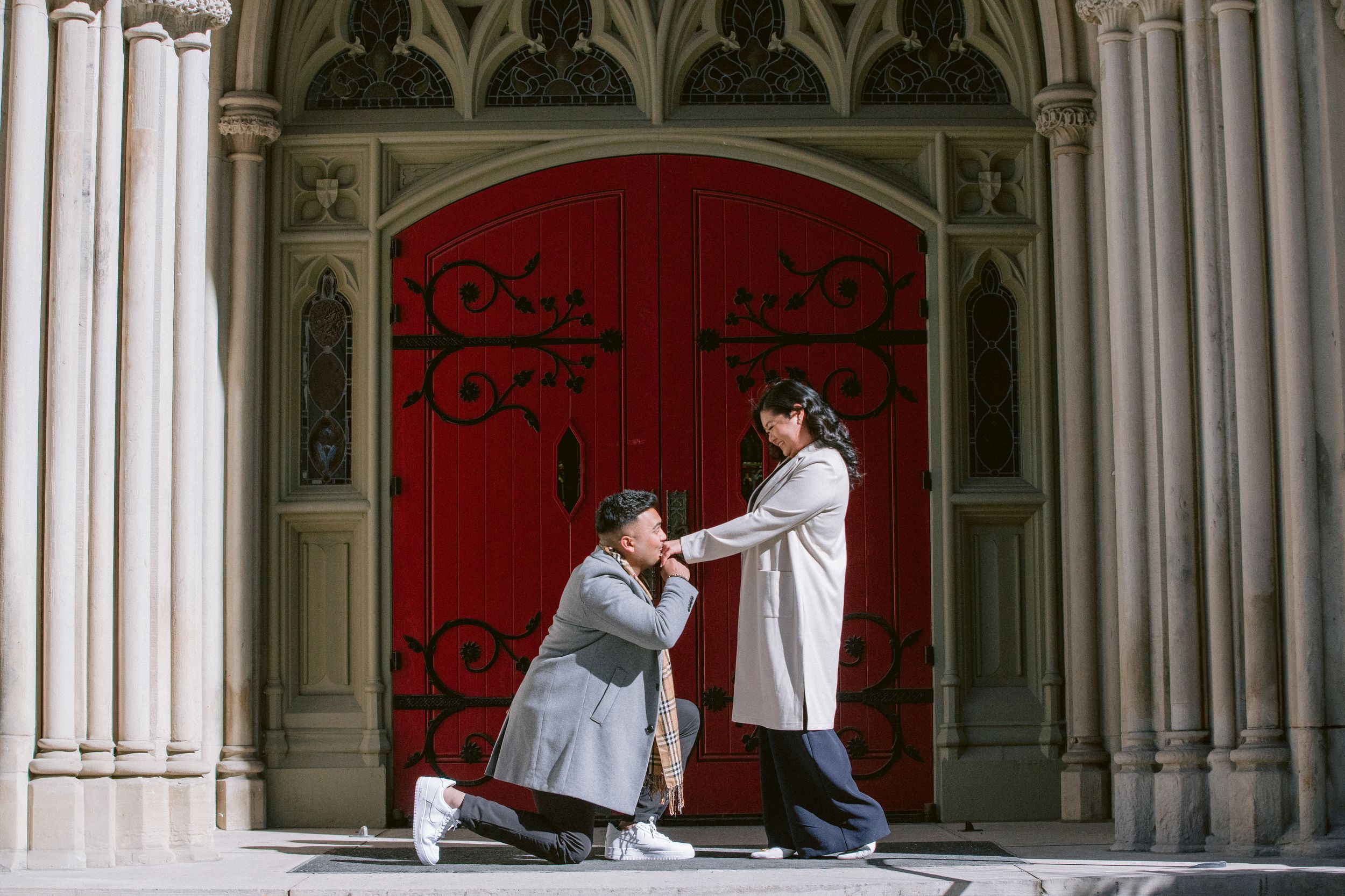 A romantic proposal scene in front of an ornate gothic church doorway with a bright red door.