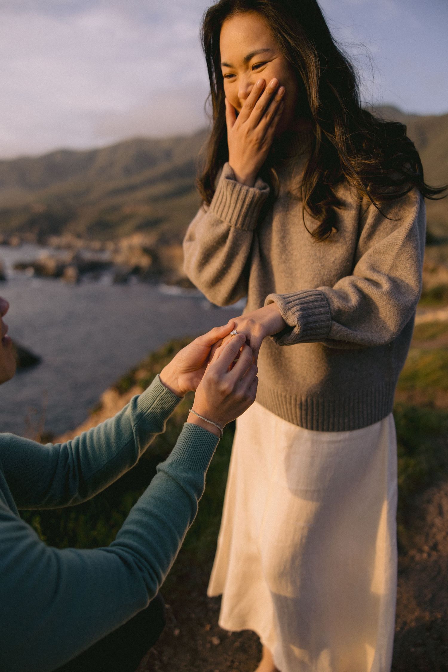 A romantic proposal moment captured during golden hour at a scenic coastal overlook with mountains in the background.