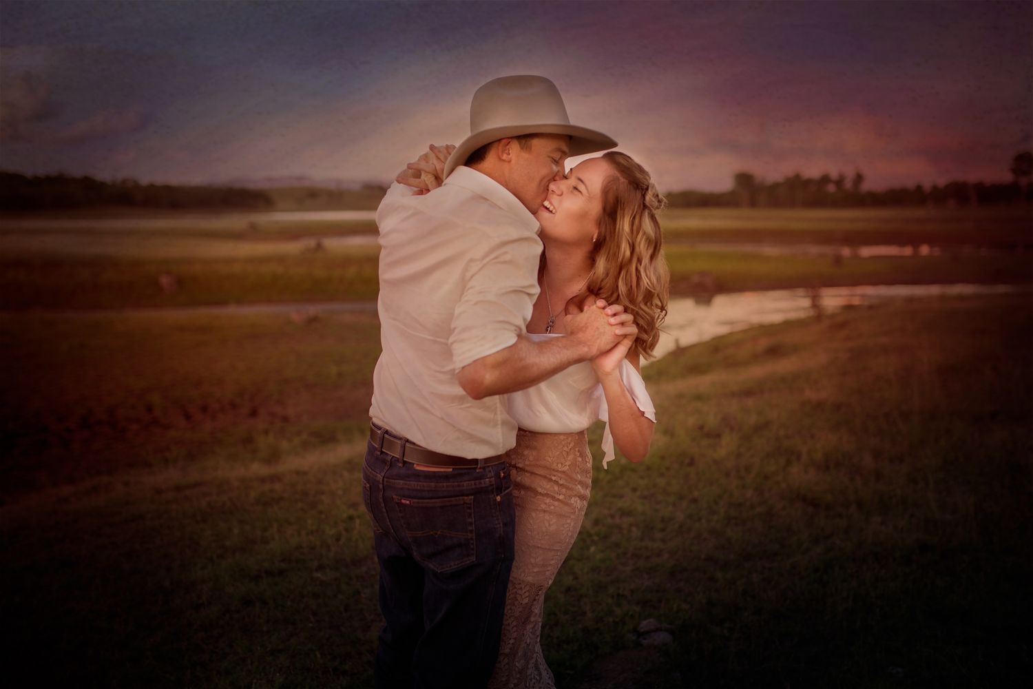 A couple embracing in a field at sunset, with the man wearing a cowboy hat and the woman looking up at him.