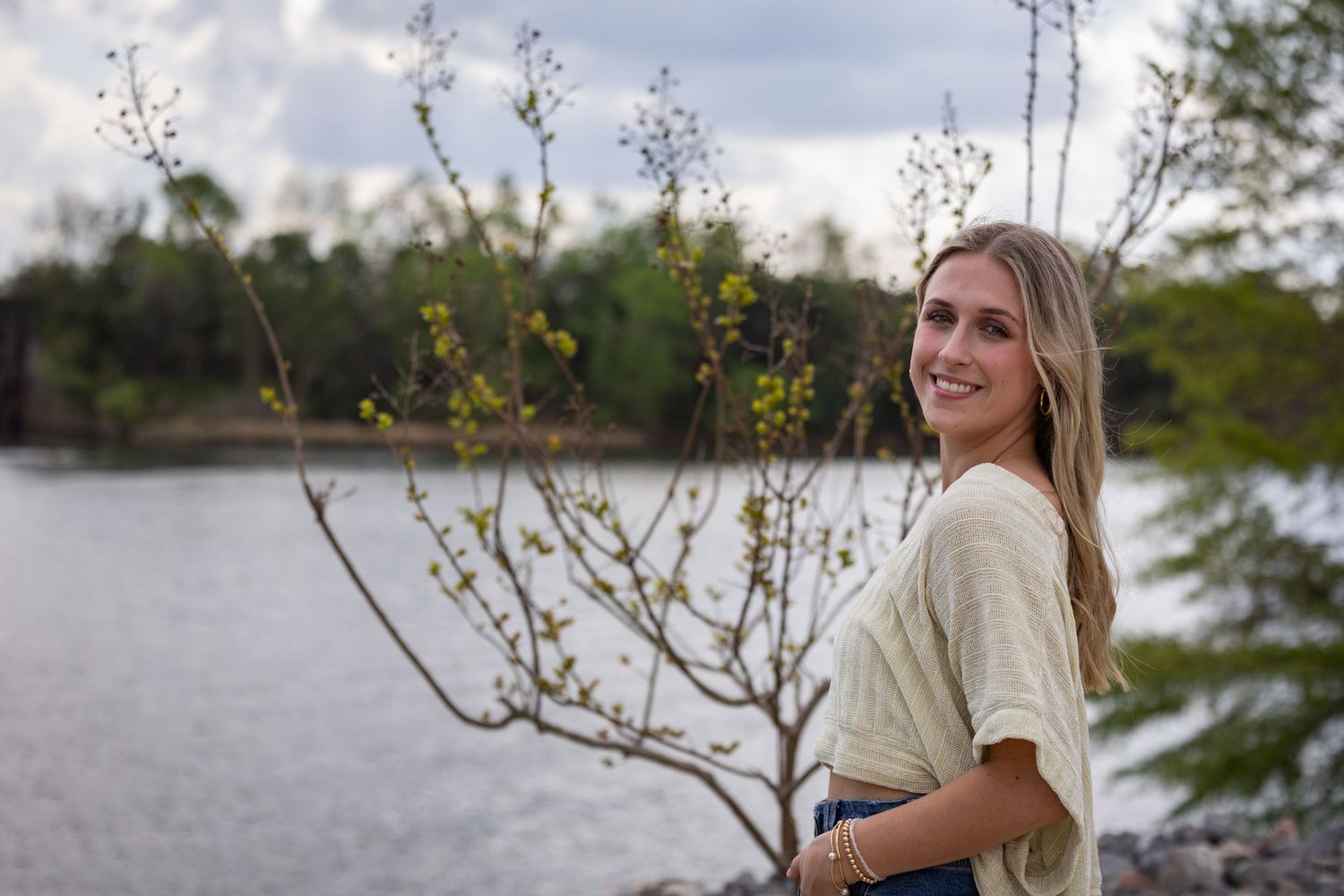 A person in casual attire stands near a lake with blooming tree branches in the foreground.