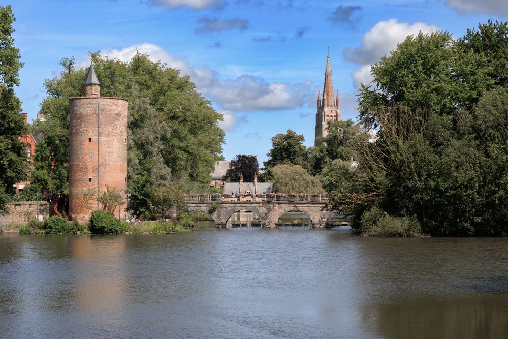 Historic tower and church spire reflect in calm waters with stone bridge in medieval English town.