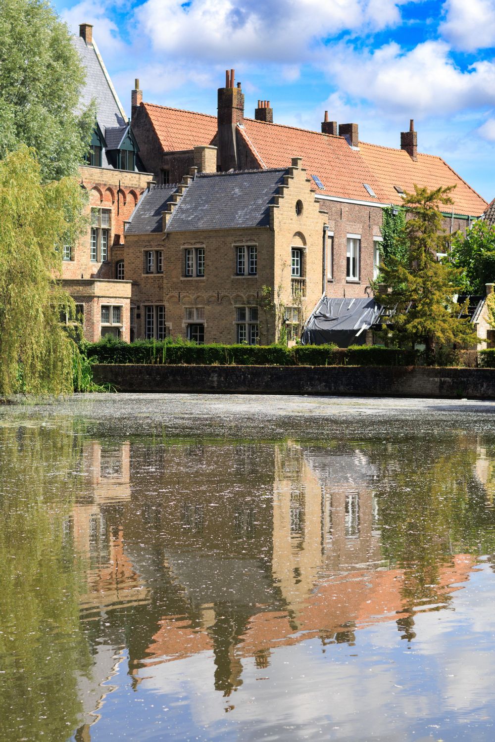 Historic brick buildings with orange tiled roofs reflect in calm canal waters of Bruges, Belgium.