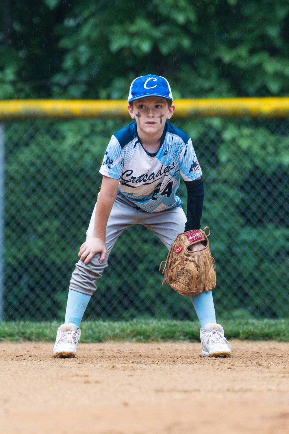 Young baseball player in light blue uniform stands ready in fielding position on dirt infield.