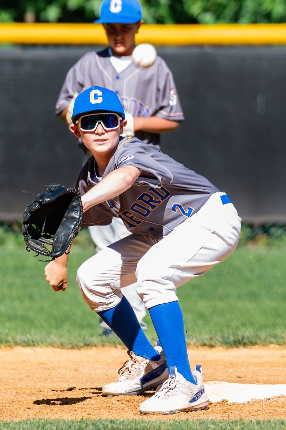 Youth baseball player in blue cap and white uniform crouches in fielding position on the diamond.