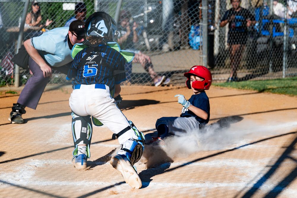 Baseball player in red slides into home plate as catcher in light blue uniform attempts to make the tag during game.