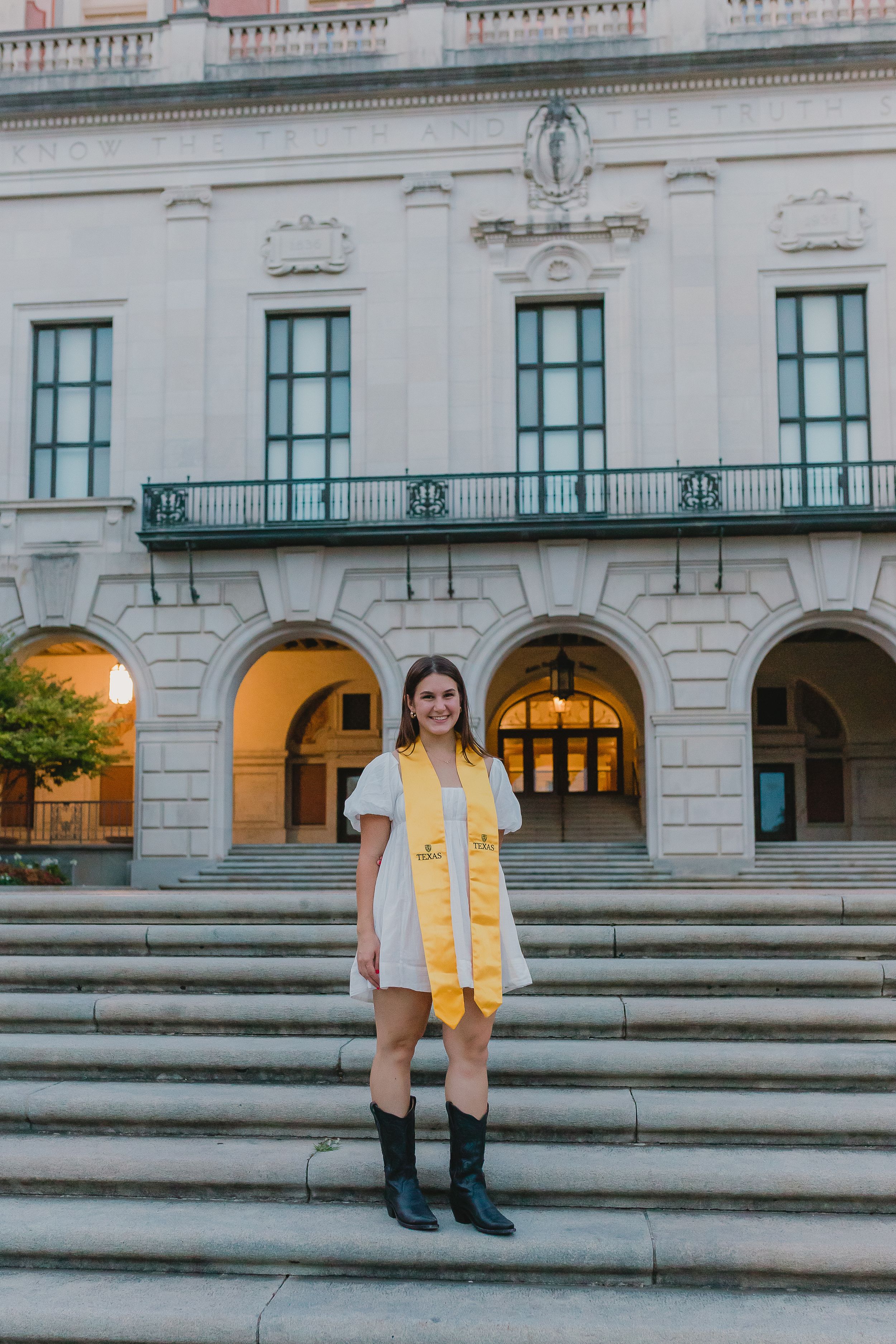 A figure wearing a yellow dress and black boots stands on stone steps in front of a historic building with arched windows.