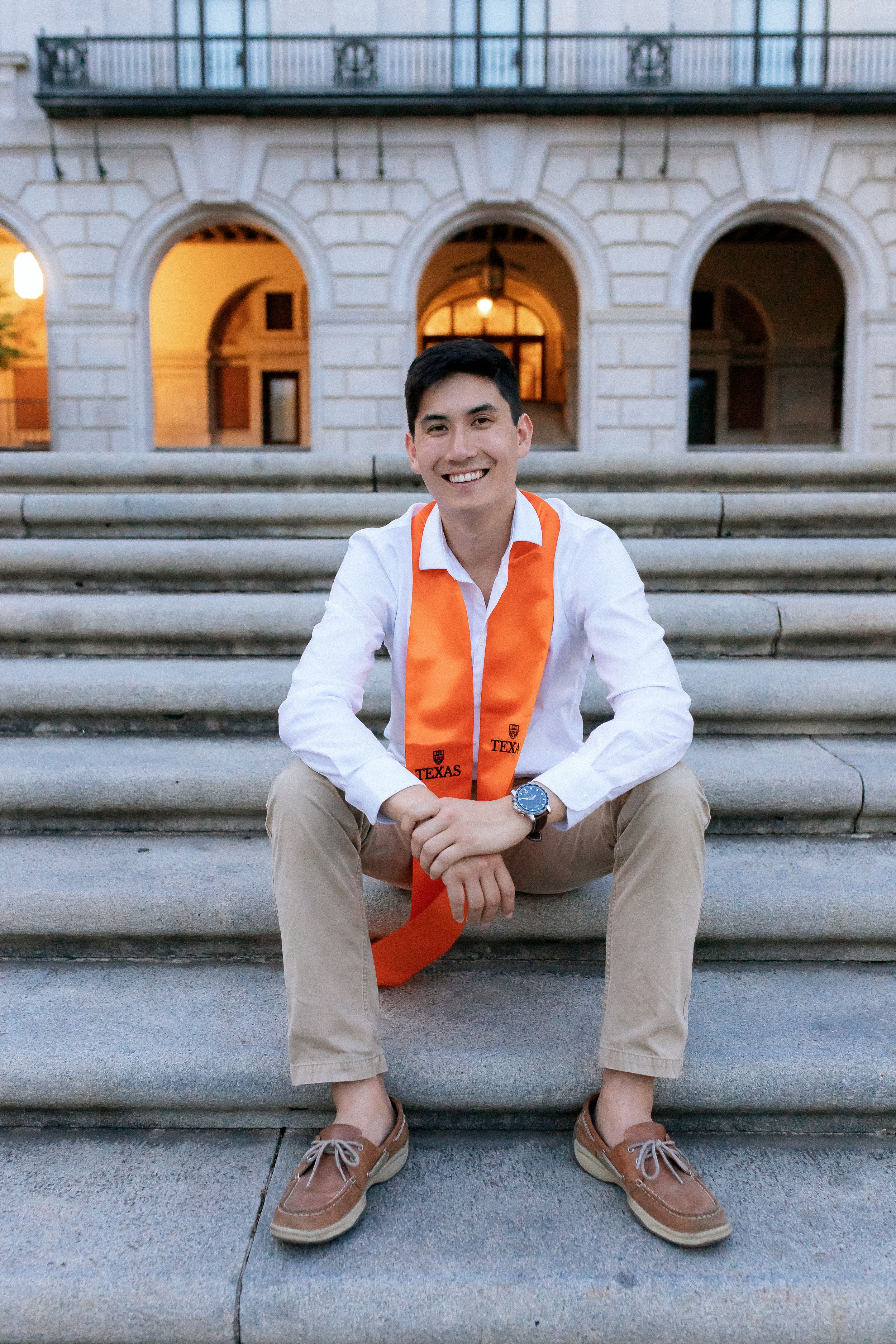 Graduate in white shirt and orange stole sitting on stone steps of historic building.