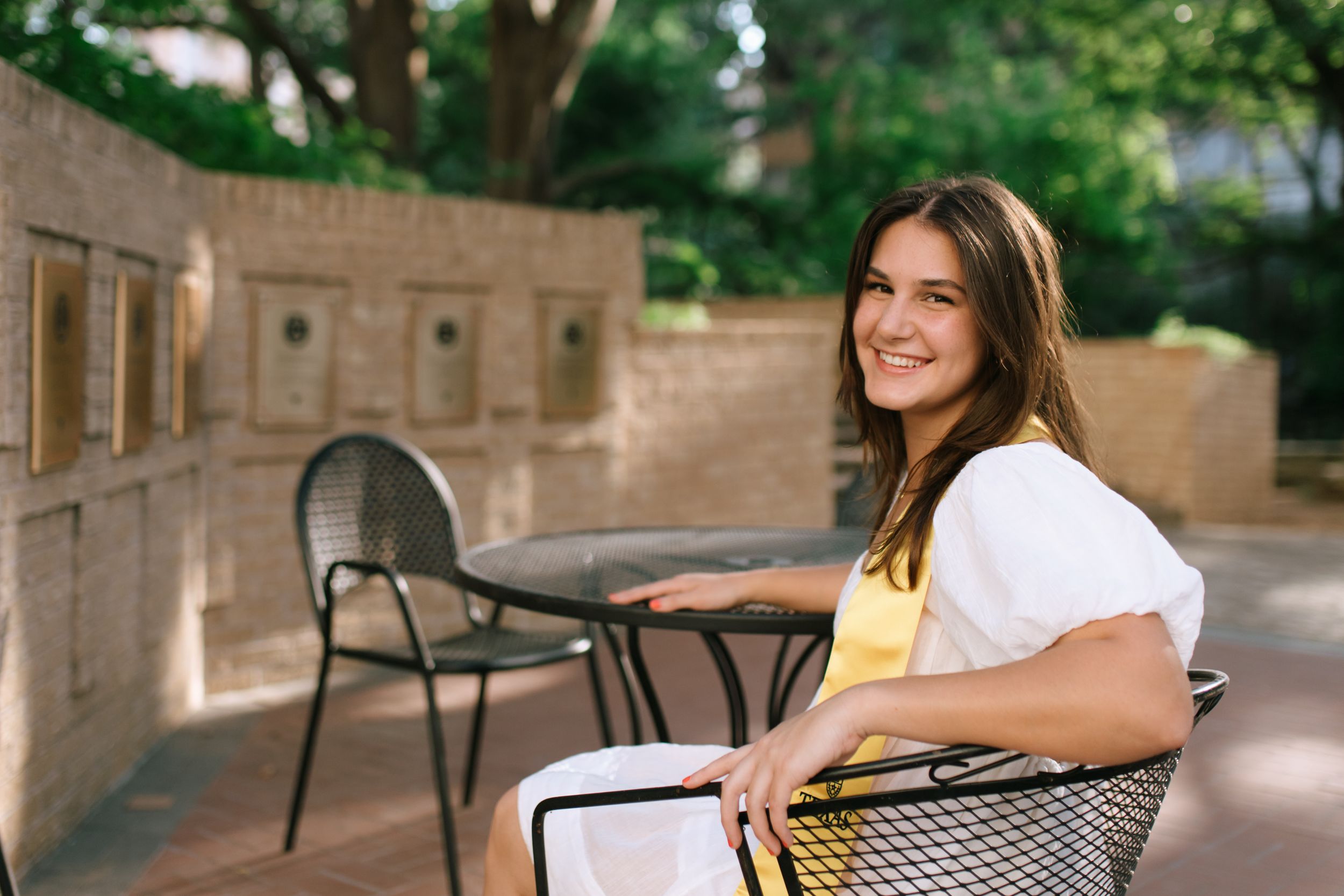 A figure sits outdoors at a metal patio chair beside a stone wall.