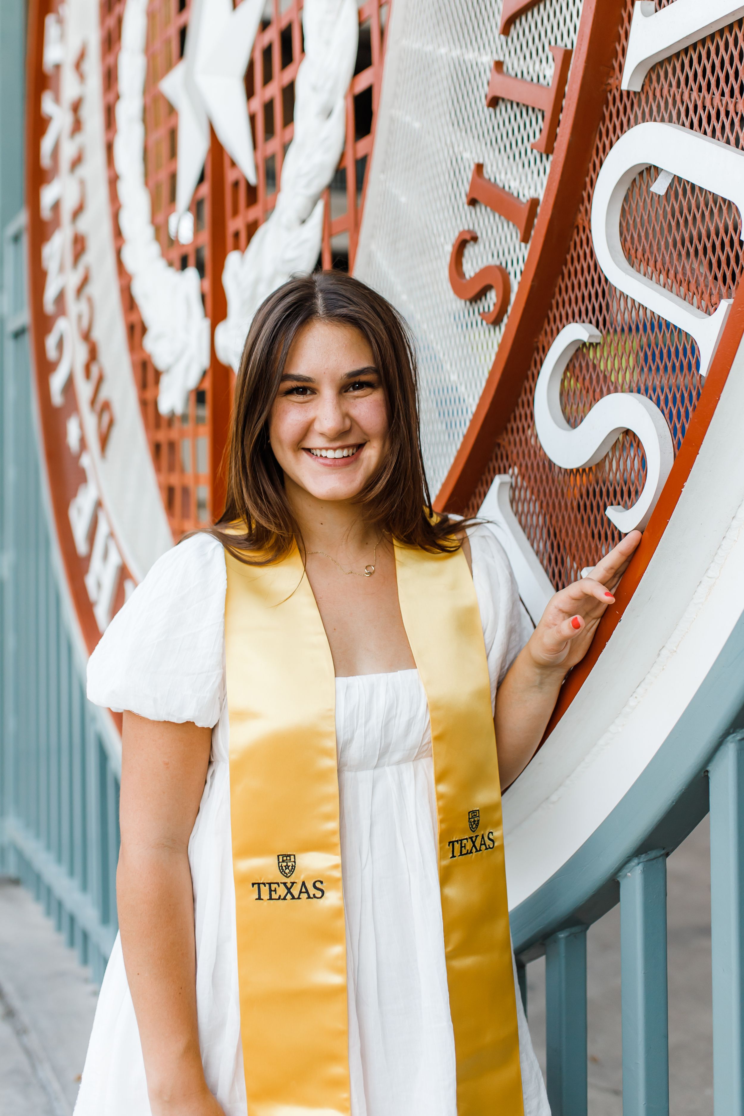 Graduate in white dress and yellow Texas stole posing next to university logo wall.