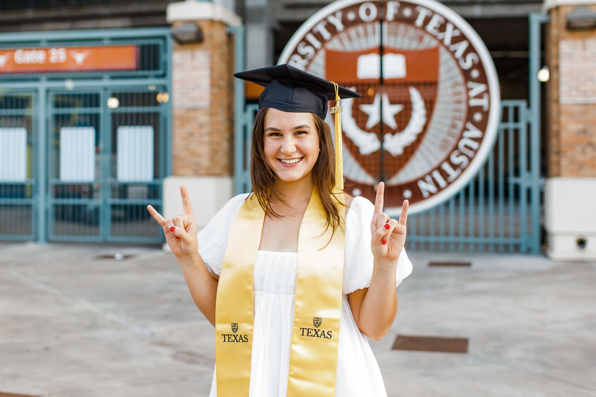 Graduate in cap and gown with yellow stole making peace signs in front of university building.