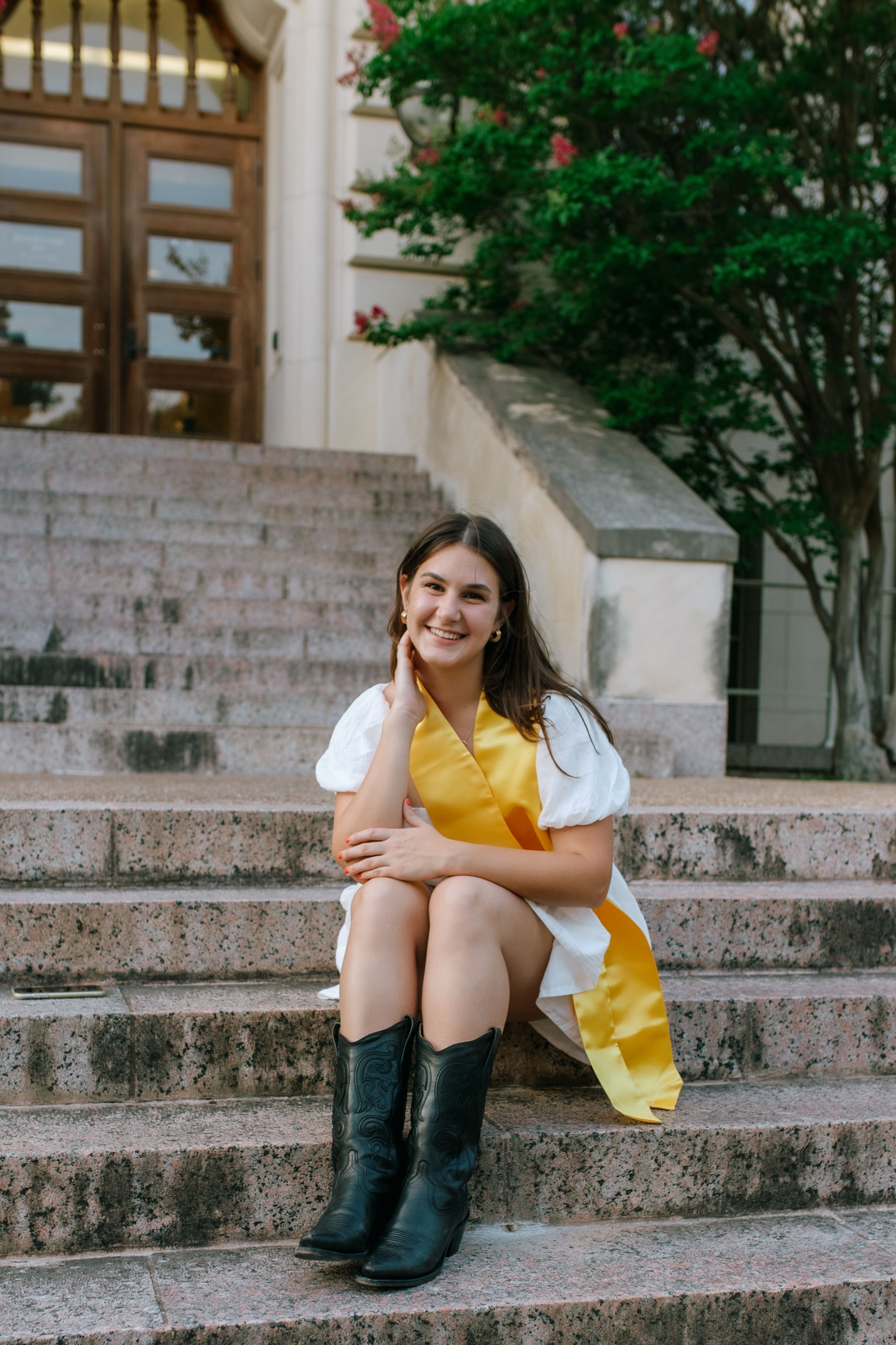 Graduate in white dress and yellow stole sitting on stone steps with boots.