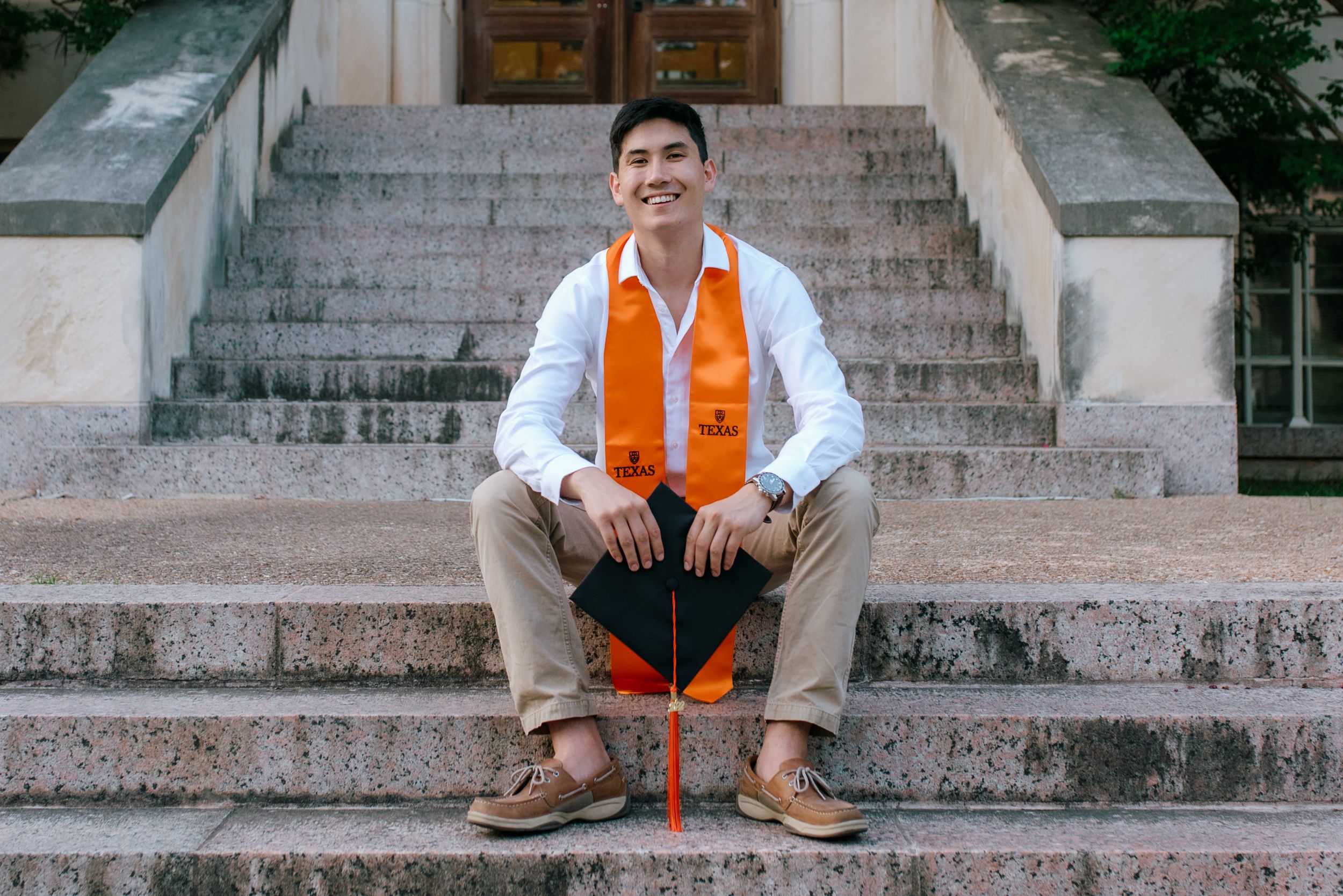 Graduate in orange stole sitting on stone steps in front of university building.