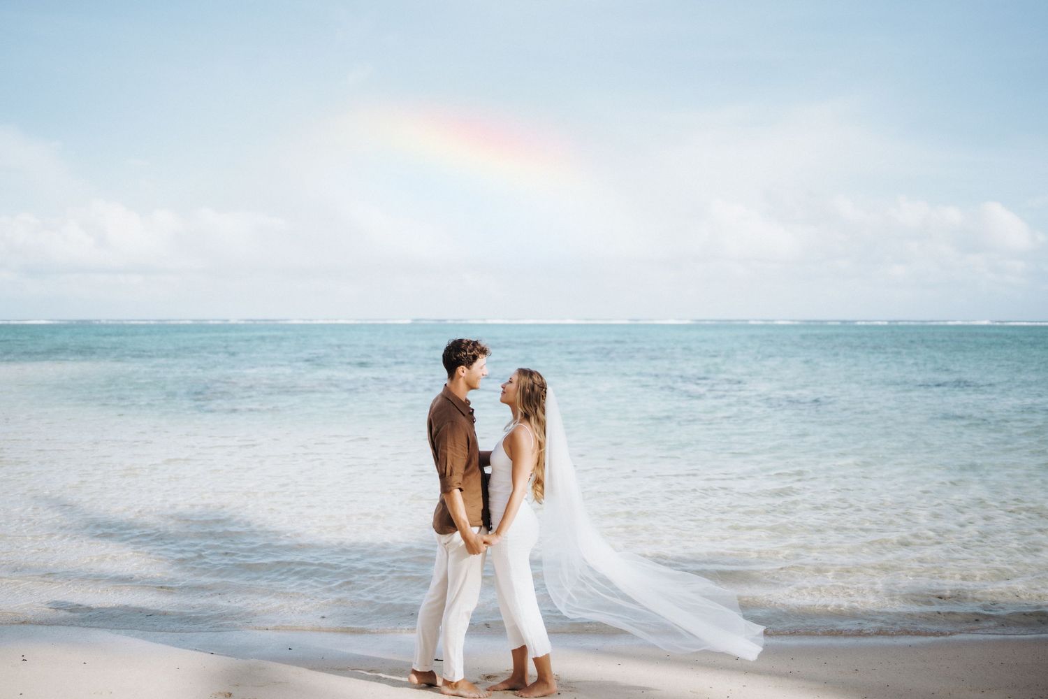 A couple dressed in white stands together on a tropical beach with a rainbow appearing in the ocean background.